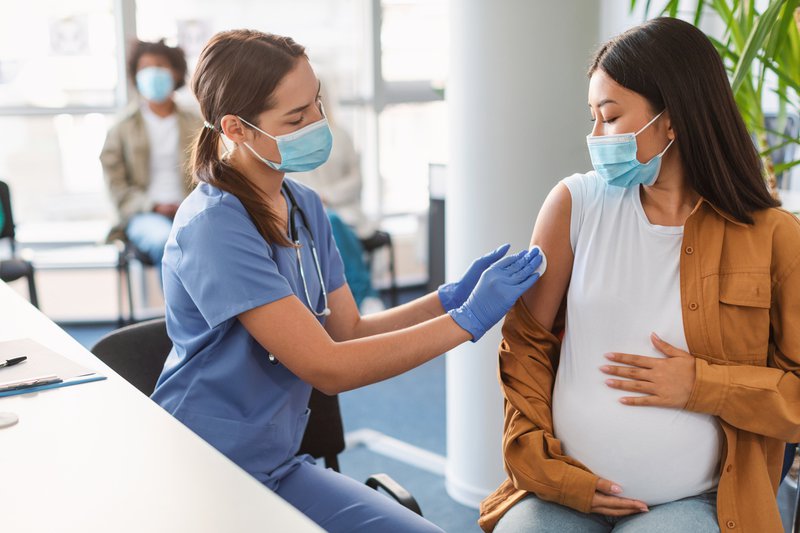 Nurse in face mask and gloves disinfecting skin of pregnant patient, rubbing shoulder with cotton pad before injection, asian lady hug belly, clinic background. Covid coronavirus treatment concept