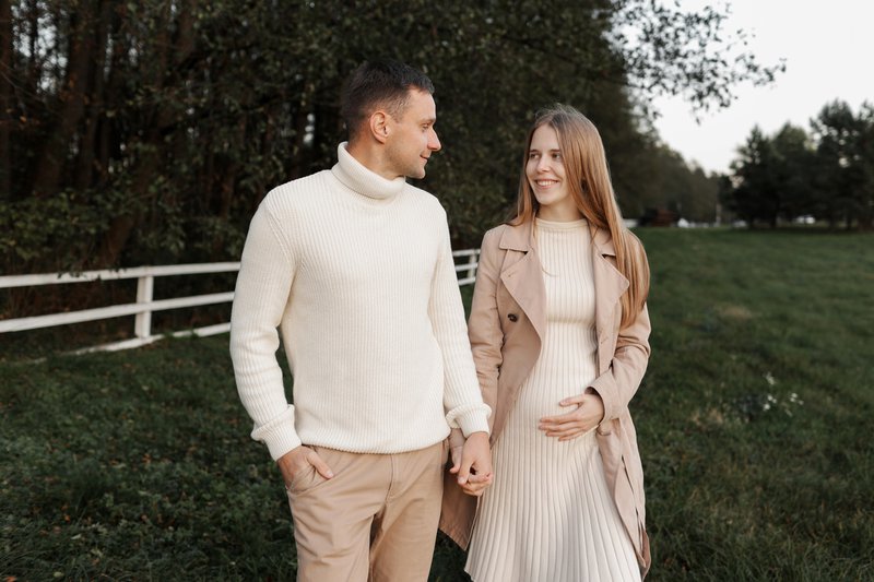 Young happy romantic pregnant couple is walking holding hands outdoors at autumn warm day. Pregnant woman in knit dress and her handsome man expecting a baby. Happy family day. Parenthood, motherhood.